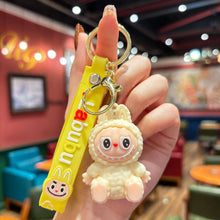 Load image into Gallery viewer, Hand holding a yellow Labubu keychain with a cartoon character in a restaurant setting
